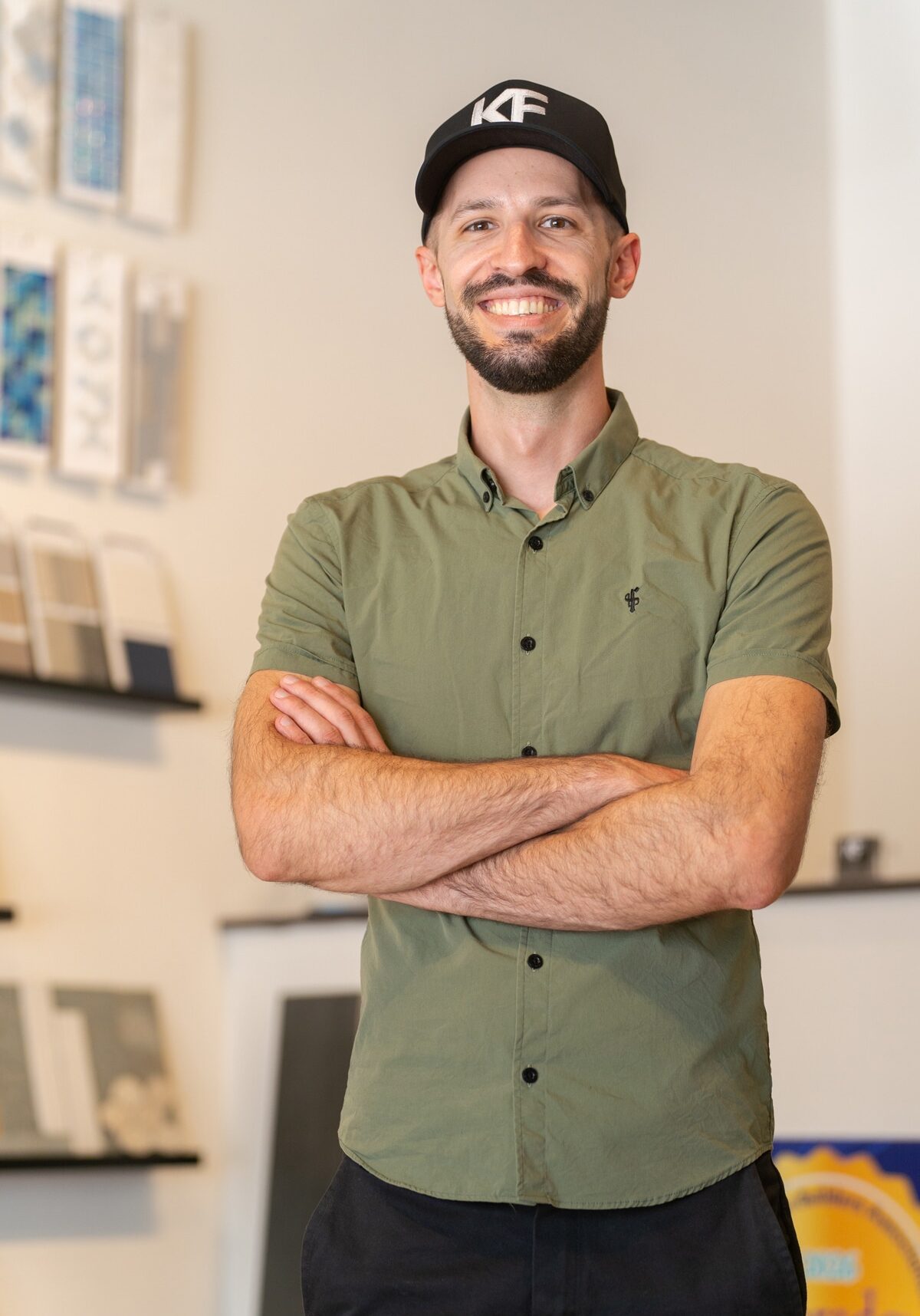 A smiling man in a green shirt and black cap stands indoors with arms crossed. Shelves with books or materials are visible in the background.