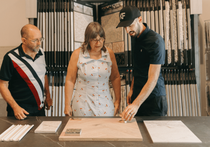 Three people stand around a table with tile samples. The man on the right points at a sample, while the other two observe. Tile displays are visible in the background.