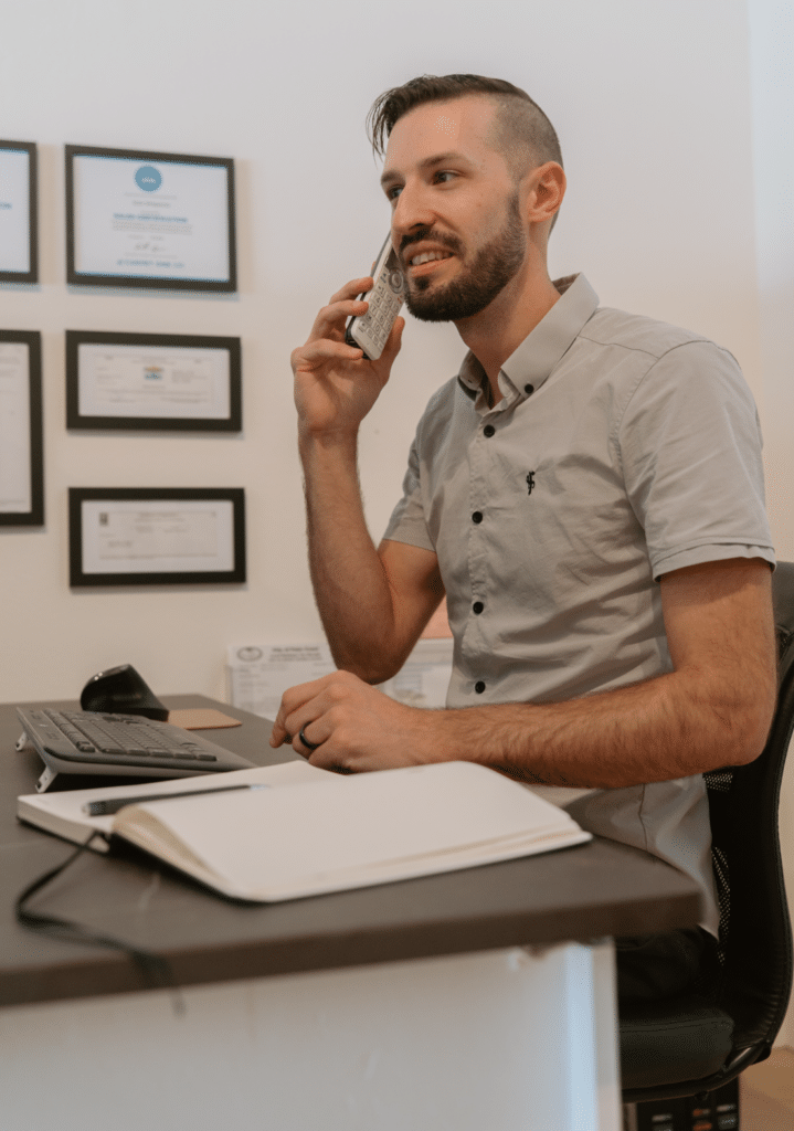 A man sits at a desk, talking on the phone and smiling. He faces a computer monitor, with framed certificates hanging on the wall behind him and a notebook on the desk in the foreground.