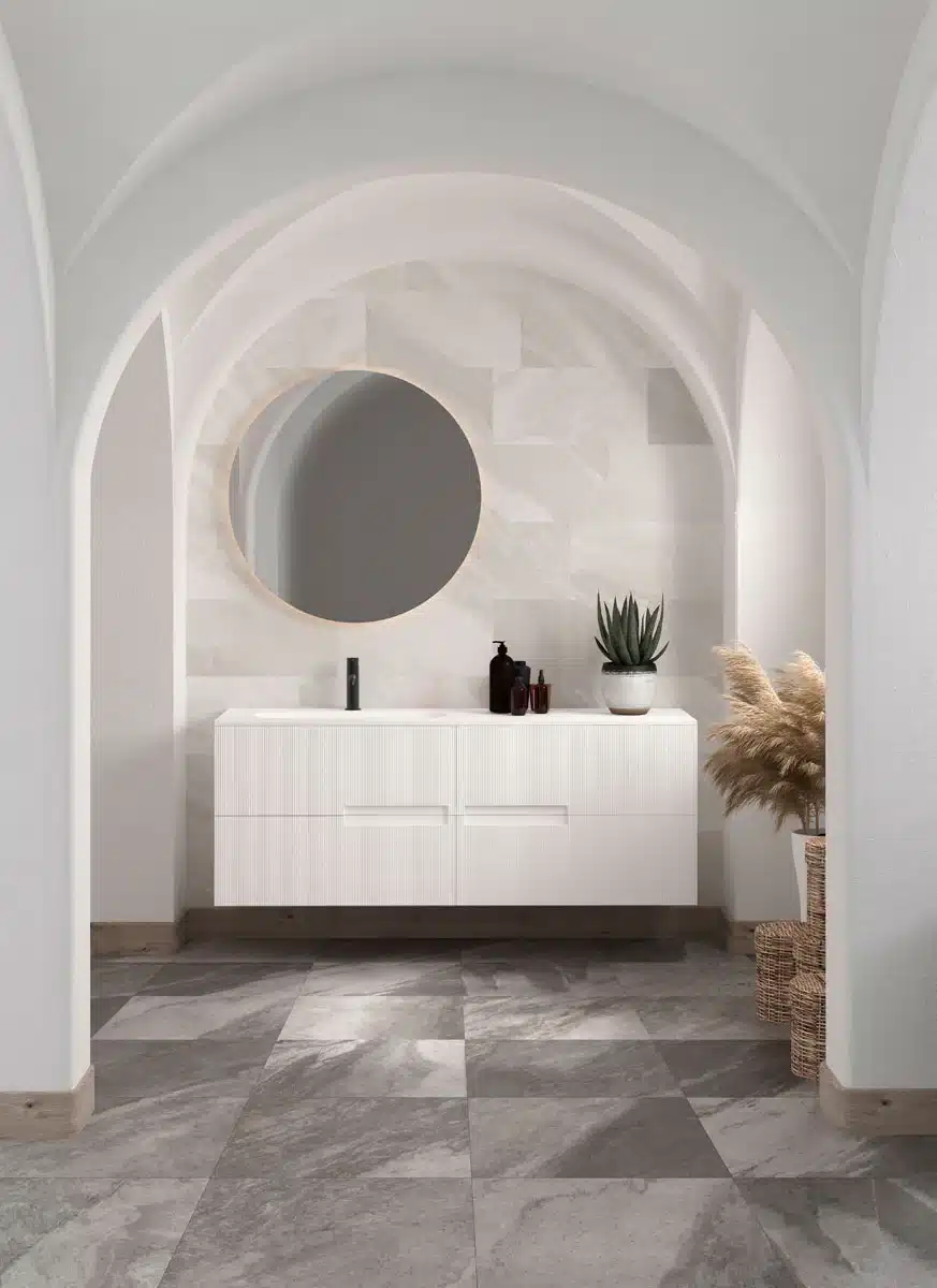 Modern bathroom with gray marble tile, a white vanity below a round mirror, a potted plant, toiletries on the countertop, and decorative pampas grass in woven baskets in the corner under arched ceilings.