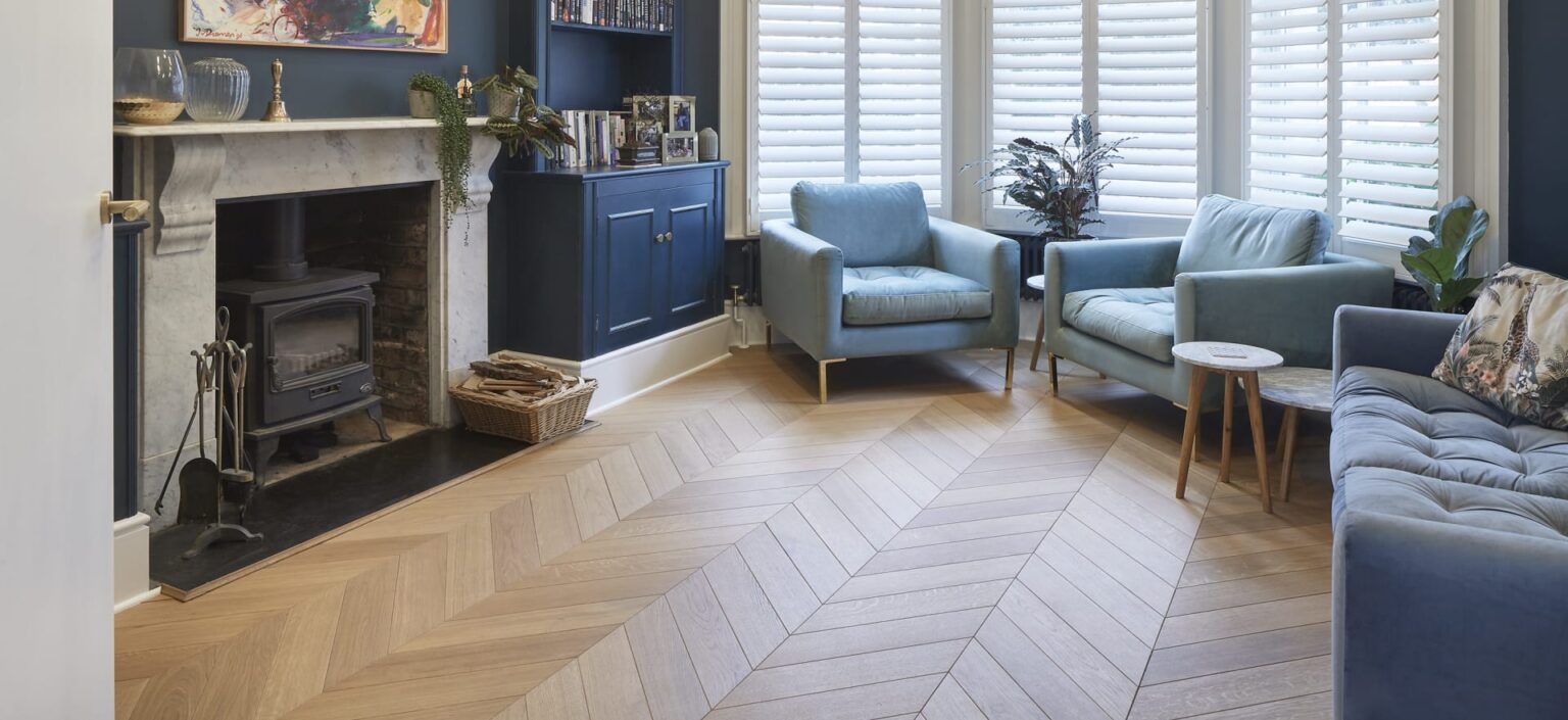 Bright living room with herringbone wood floor, blue armchairs, a blue sofa, white shutters, fireplace with wood burner, plants, and built-in blue cabinets.