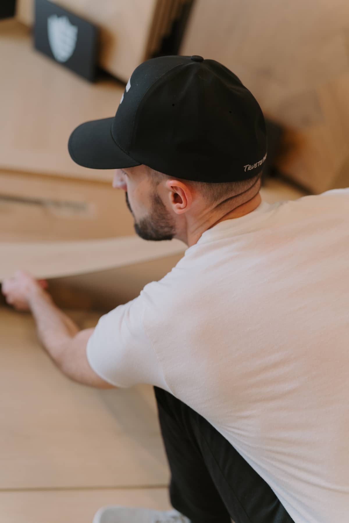 A person wearing a black cap and white t-shirt is crouching and handling a large wooden board in an indoor setting.