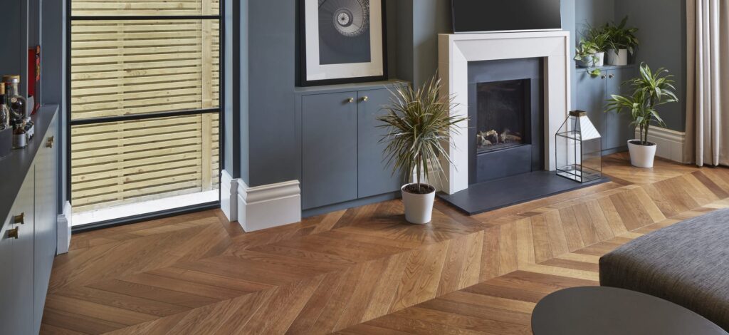 Modern living room with herringbone wood floor, blue cabinets, a fireplace, potted plants, a large framed print, and a glass door revealing a wooden fence outside.