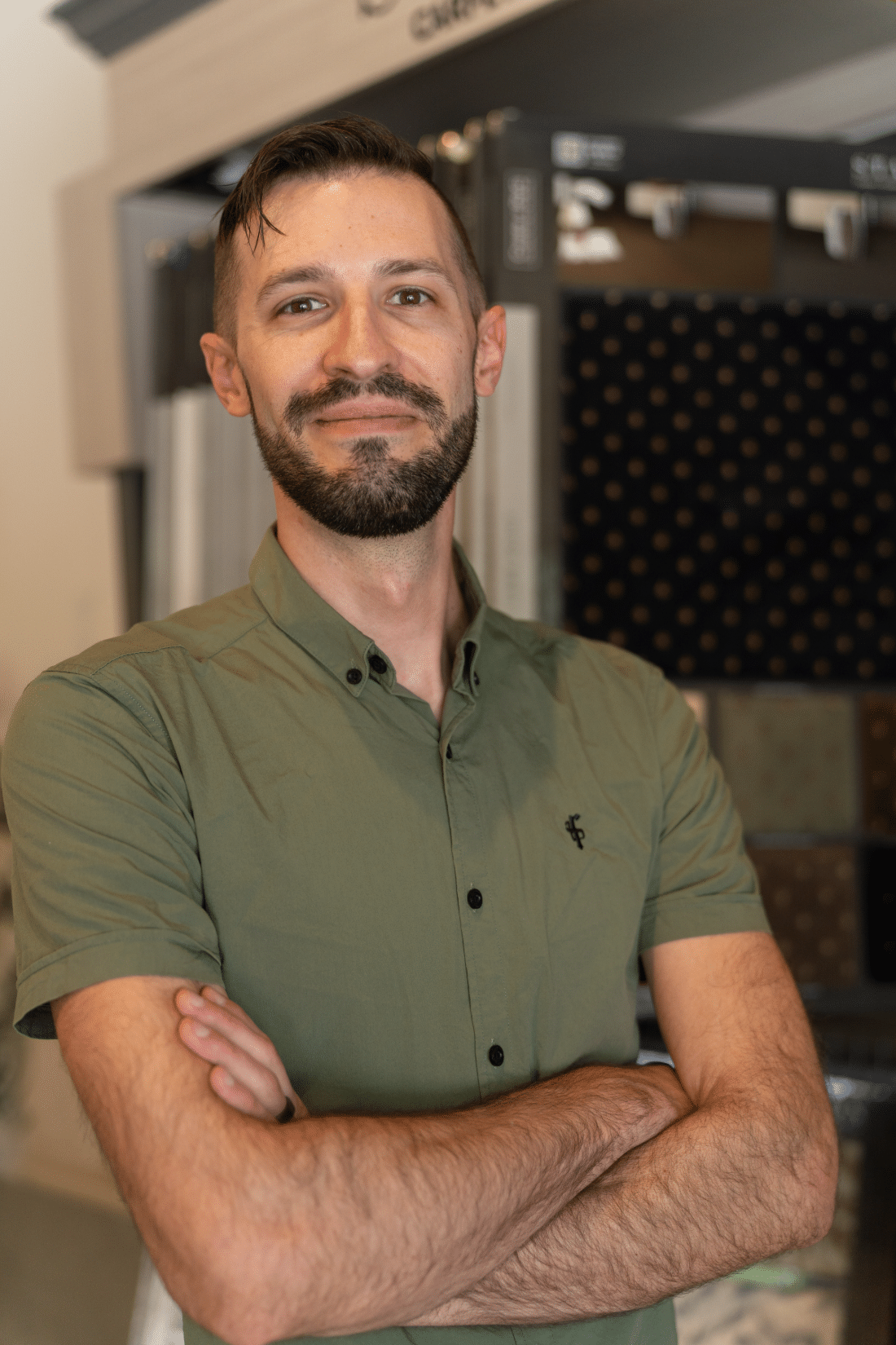 A man with short brown hair and a beard, wearing a green button-up shirt, stands indoors with arms crossed and smiles at the camera. Fabric samples are displayed in the background.