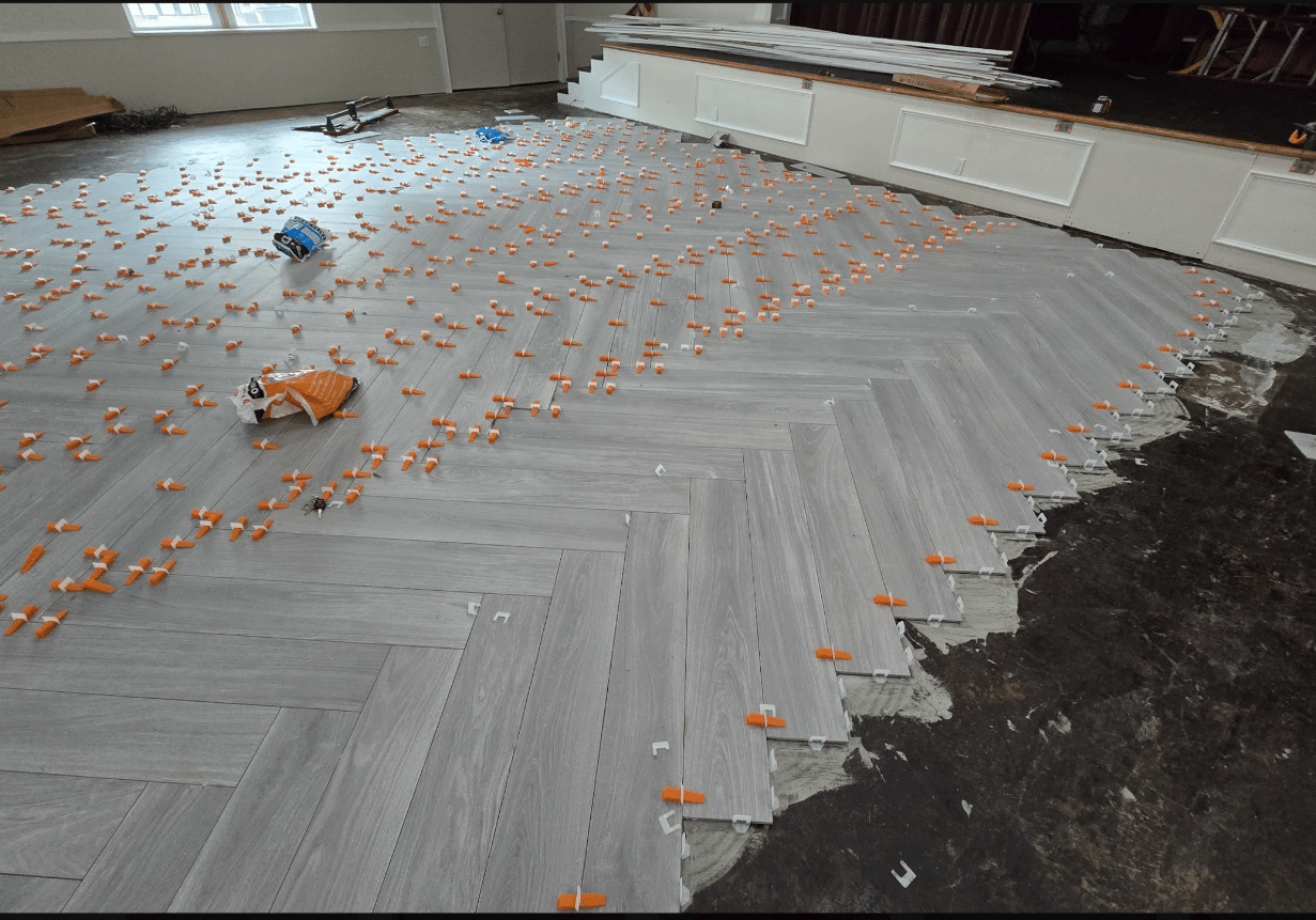 Herringbone-patterned light gray tiles being installed on a floor, with spacers and clips visible. The job is incomplete, and supplies are scattered on the tiles. The unfinished edge reveals adhesive and bare floor.