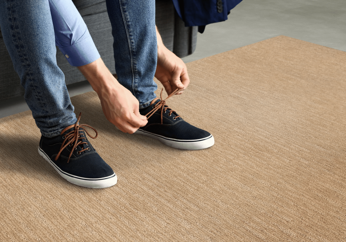 A person wearing jeans and a blue shirt ties the laces of their navy sneakers while sitting on a gray couch, with a beige rug beneath their feet.