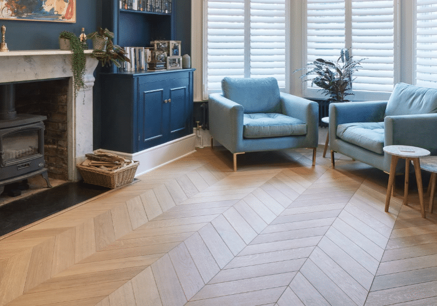 A cozy living room with blue armchairs, a matching sofa, a wood-burning stove, herringbone wood floor, houseplants, and large windows with white shutters.