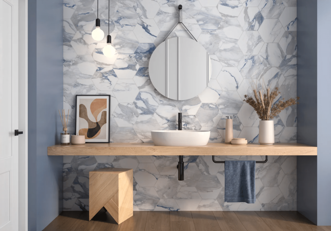 A modern bathroom features a wooden vanity with a white vessel sink, a circular mirror, and blue-and-white marble hexagonal wall tiles. The decor includes pendant lights, a geometric wooden stool, a framed abstract print, and dried flowers in a vase.