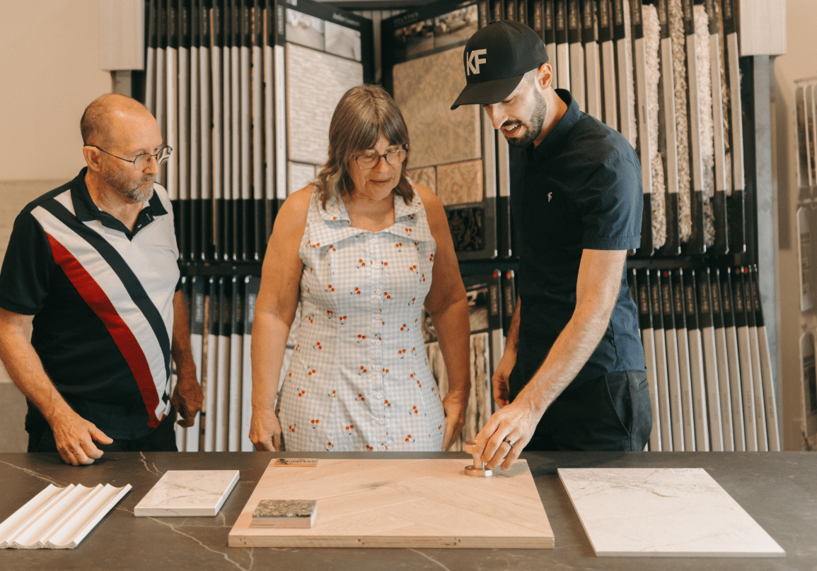 Three adults stand around a table looking at tile samples. A man in a black shirt and cap gestures toward a large tile, while the other two, an older man and woman, observe and discuss the options.